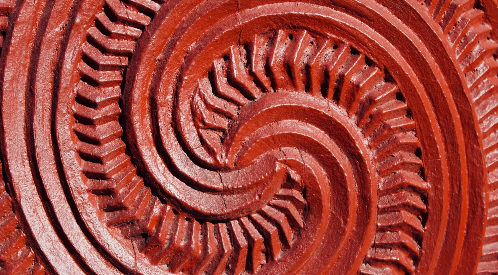 Close-up of a double koru spiral carved into wood and painted with red kōkōwai ochre