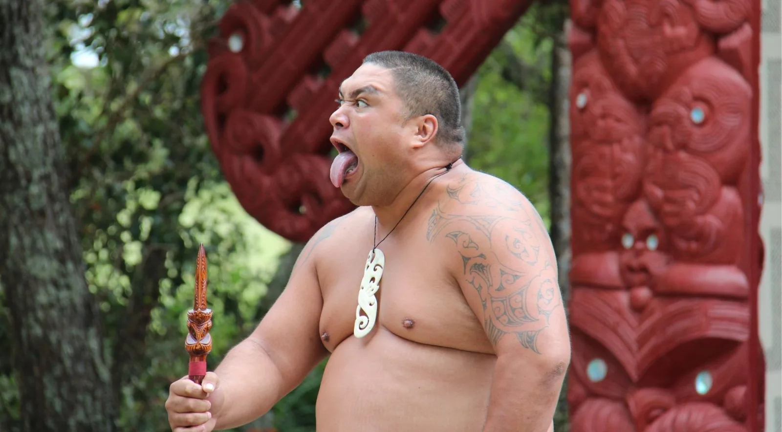 Māori warrior holding a carved taiaha, tongue extended in the wero challenge, in front of a red carved meeting house