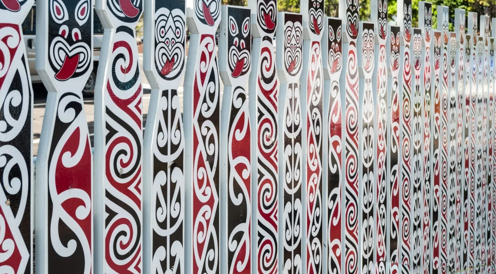 Row of tall carved fence palings painted with red, white and black koru and face designs