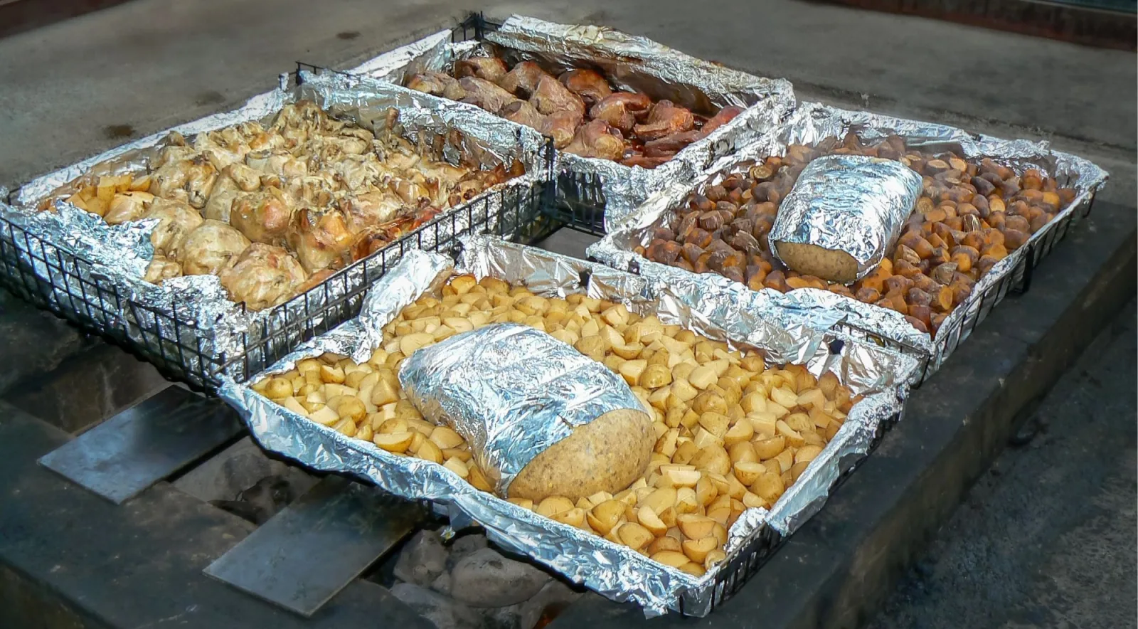 Foil-lined trays of hāngī meat, potatoes and kūmara in wire baskets, lifted from the earth oven