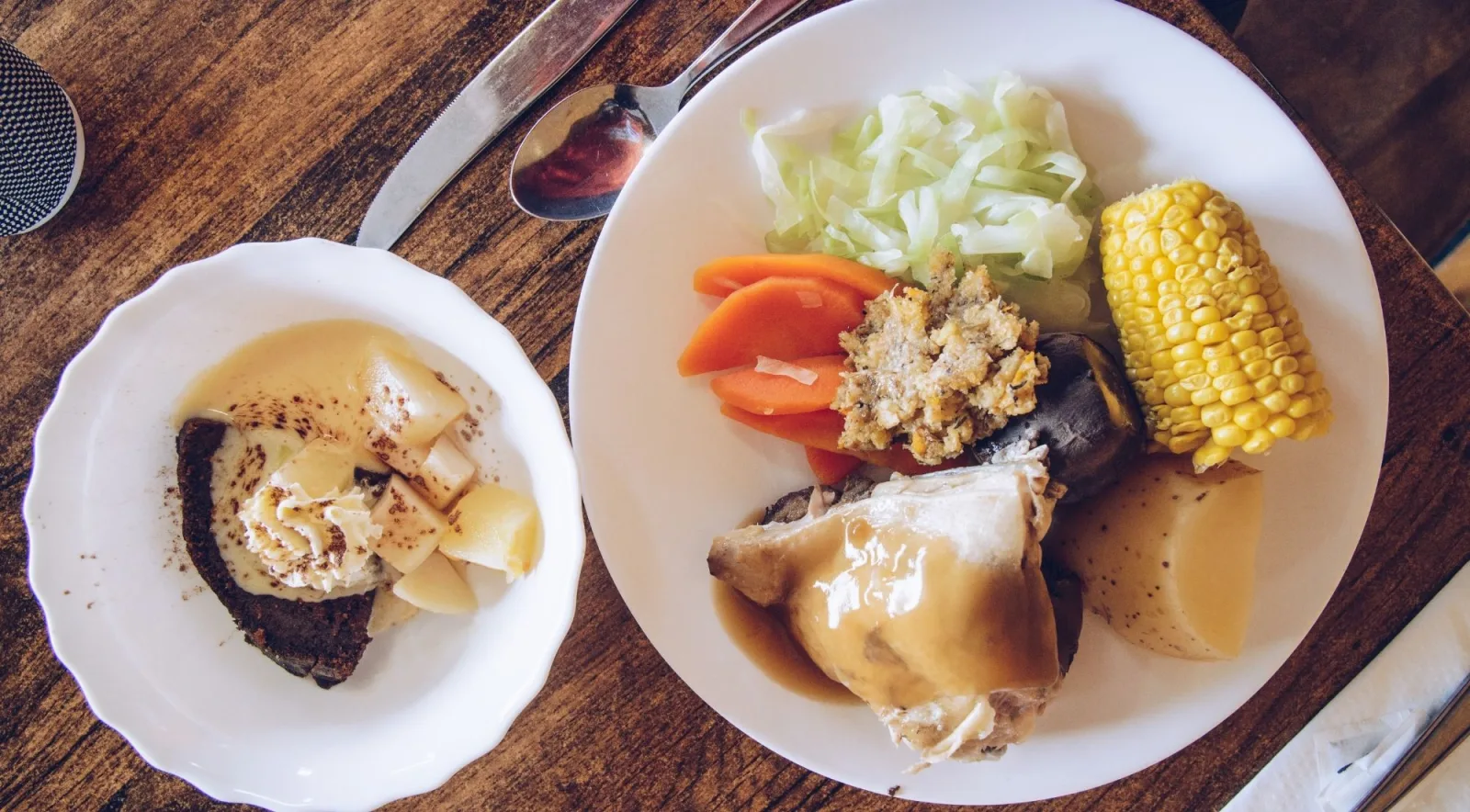 Plate of hāngī food — chicken in gravy, kūmara, potato, corn, carrot and cabbage — with a steamed pudding alongside