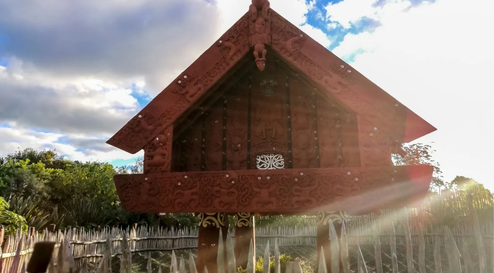 Carved red pātaka storehouse behind a wooden palisade of sharpened stakes, backlit by low sun