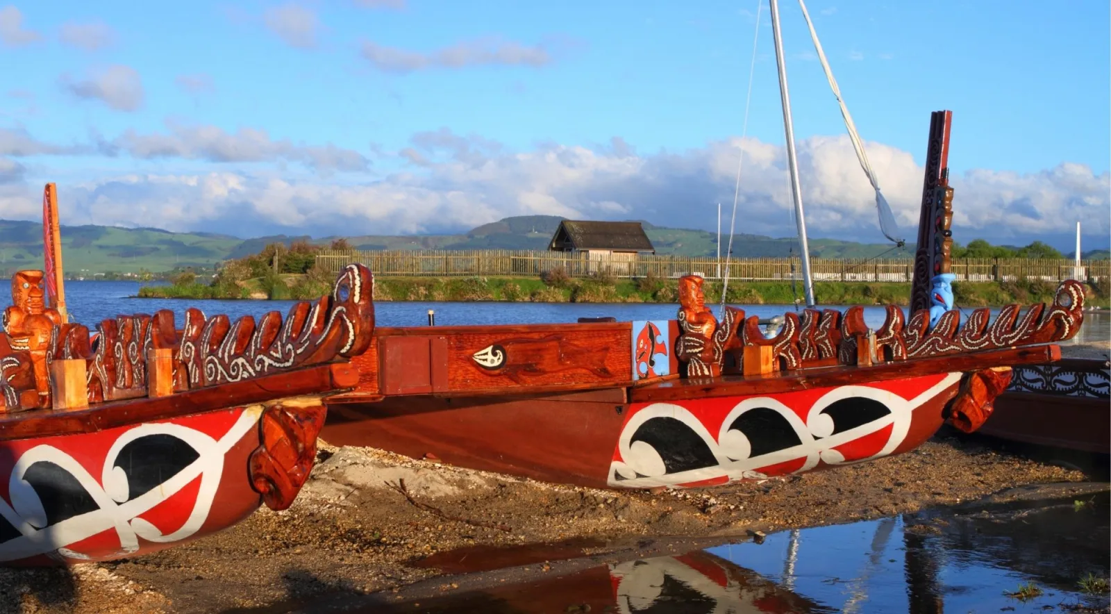 Two carved waka taua drawn up on the shore of Lake Rotorua with green hills across the water