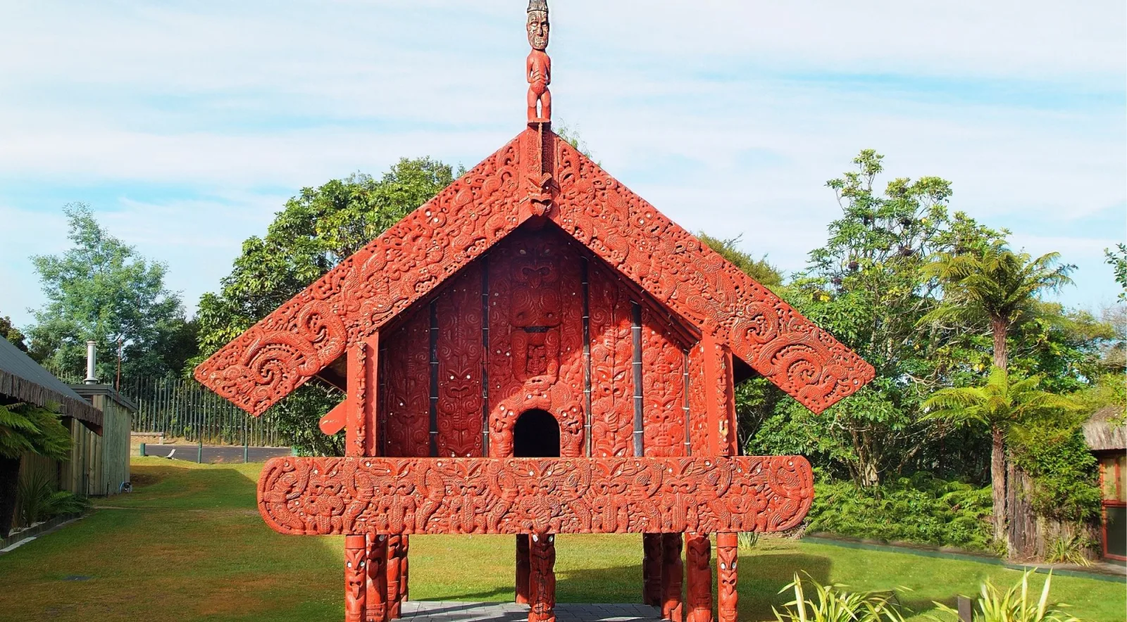 Ornately carved red pātaka storehouse raised on carved posts, surrounded by lawn and tree ferns