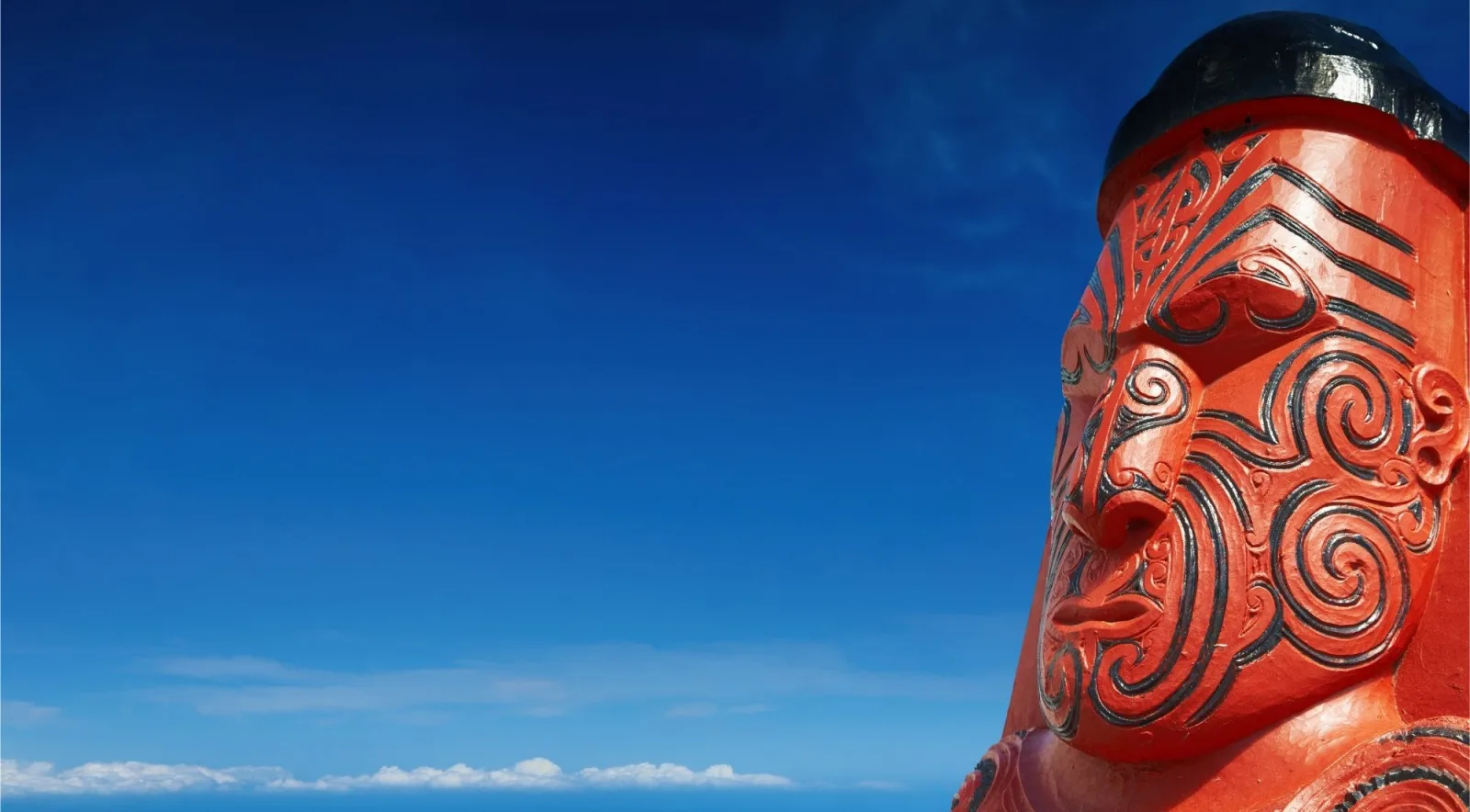 Close-up of a red carved head with black spiral moko patterns, set against a deep blue sky
