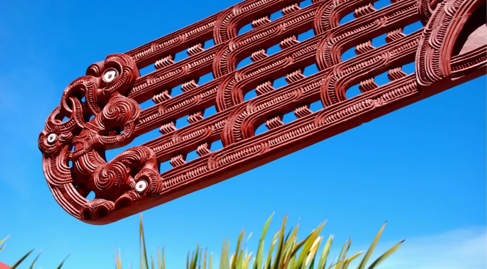 Carved red maihi bargeboard of a meeting house angled against a blue sky, with flax leaves below