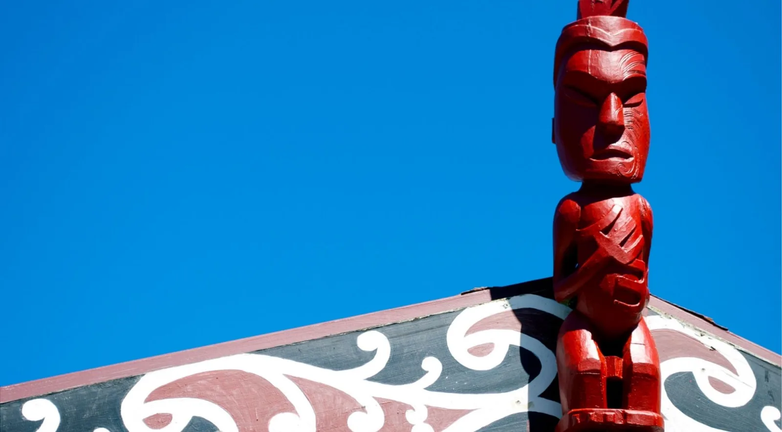 Red carved tekoteko figure standing on the gable of a meeting house against a clear blue sky