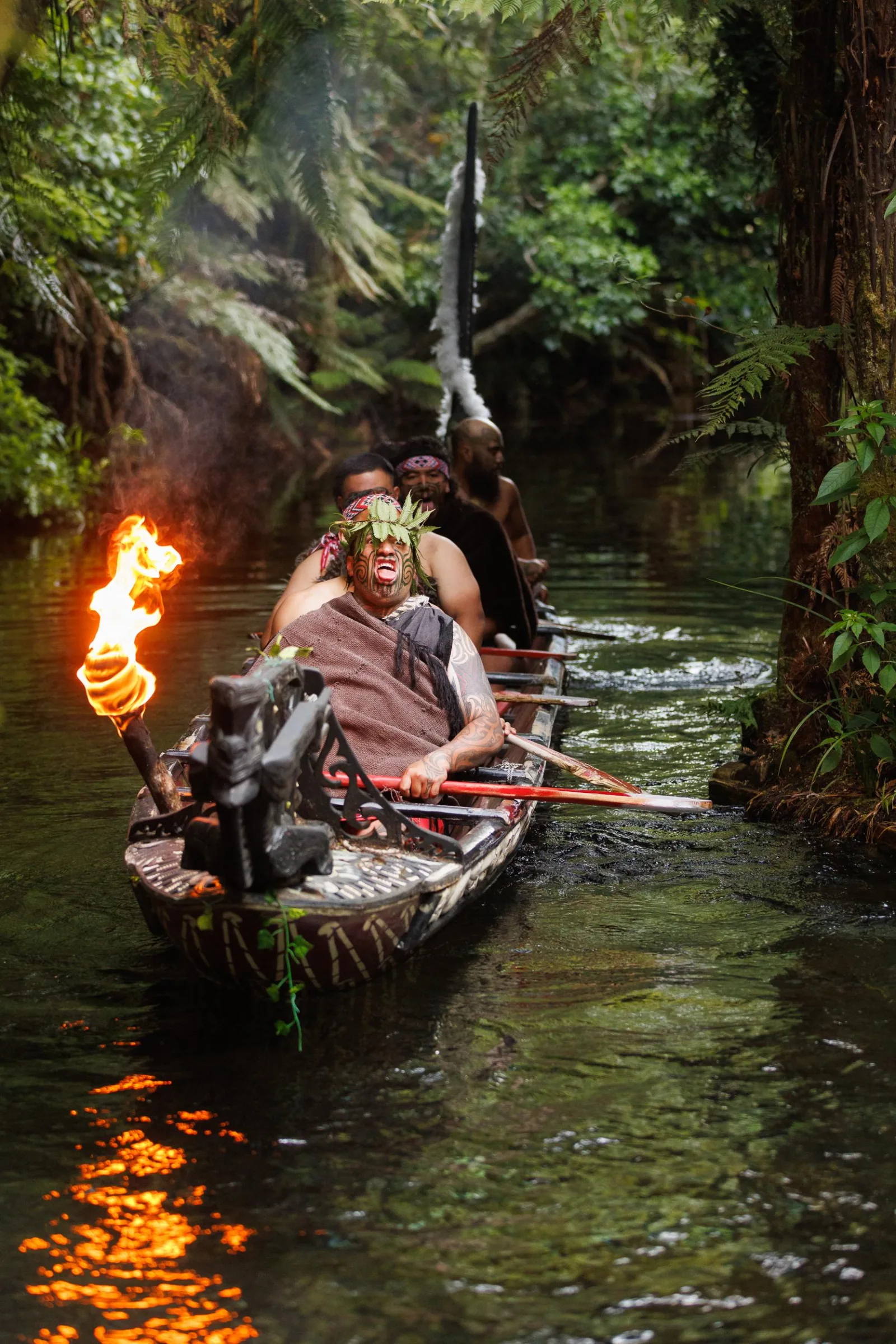 A chanting warrior leading a carved waka down the Wai-o-whiro stream by torchlight at Mitai Māori Village