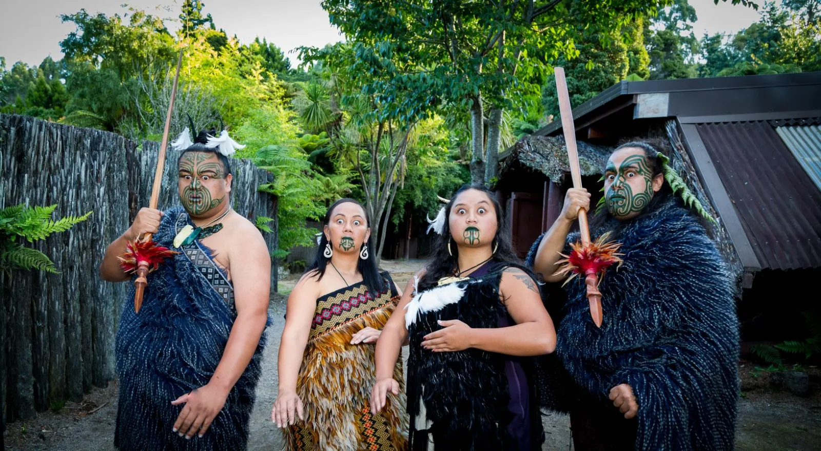Four Mitai performers in feather kākahu cloaks with moko, standing by a palisade and thatched whare