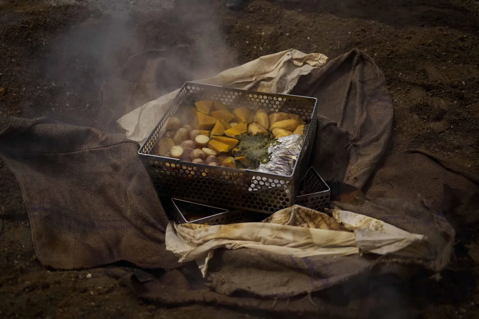 A steaming hāngī basket of kūmara and potato lifted onto hessian sacking beside the pit