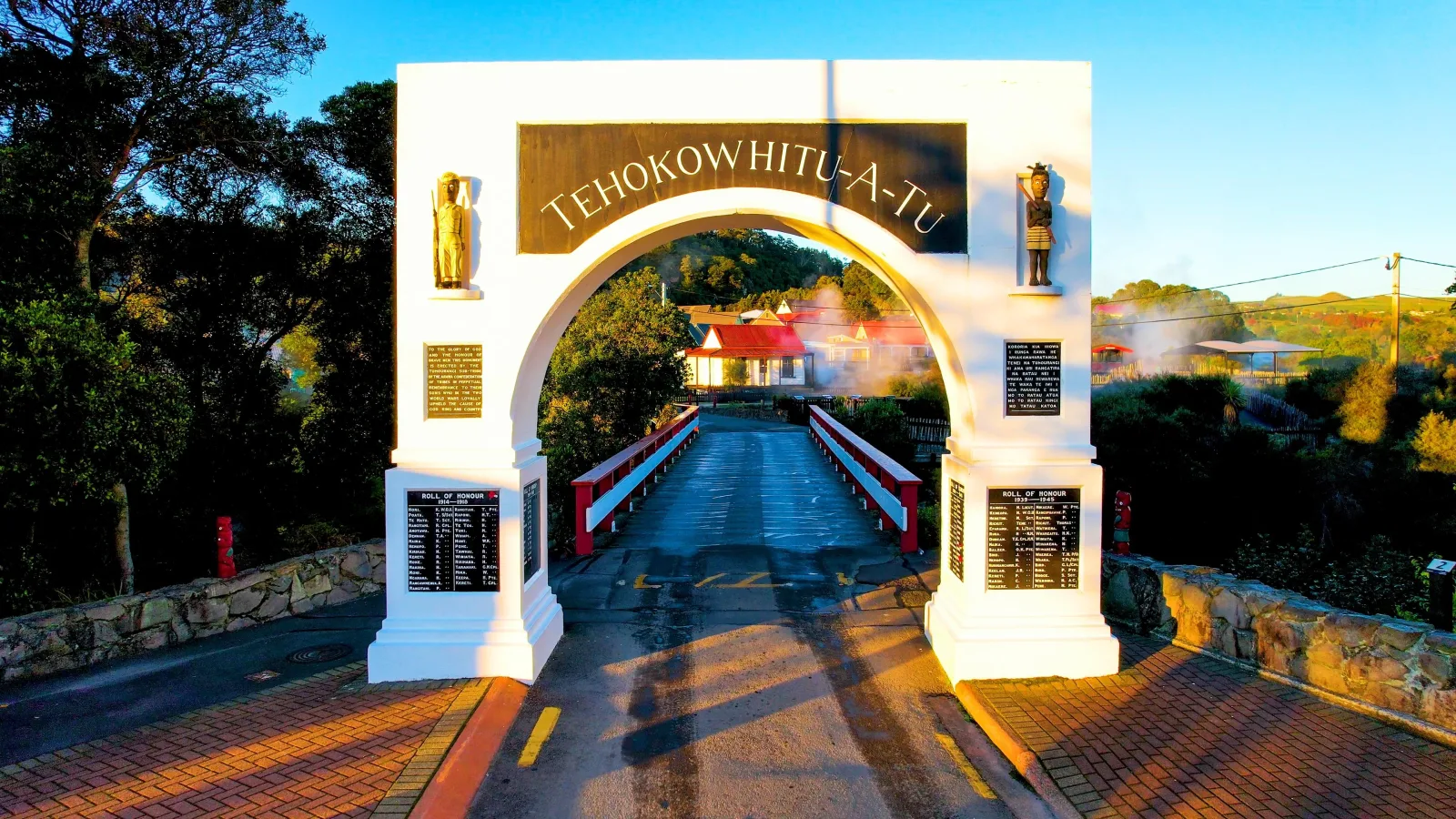 Te Hokowhitu-a-Tū memorial archway at Whakarewarewa, with the village houses and rising steam beyond it
