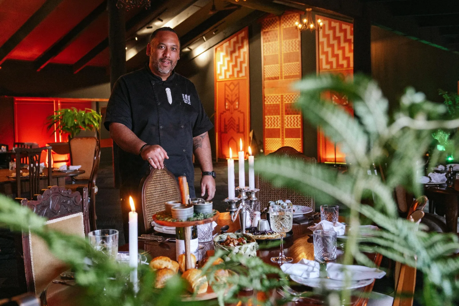 A Te Pā Tū chef at a candlelit dining table set for the four-course menu, tukutuku panels lit behind