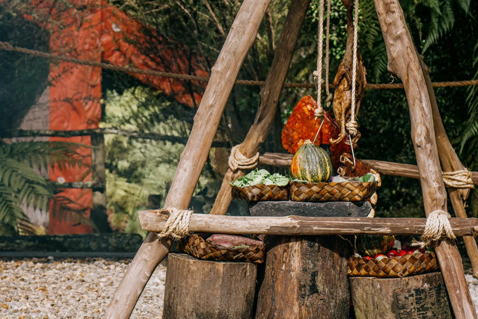 Woven kete of seasonal produce — squash, greens and meat — hung on a wooden frame at Te Pā Tū