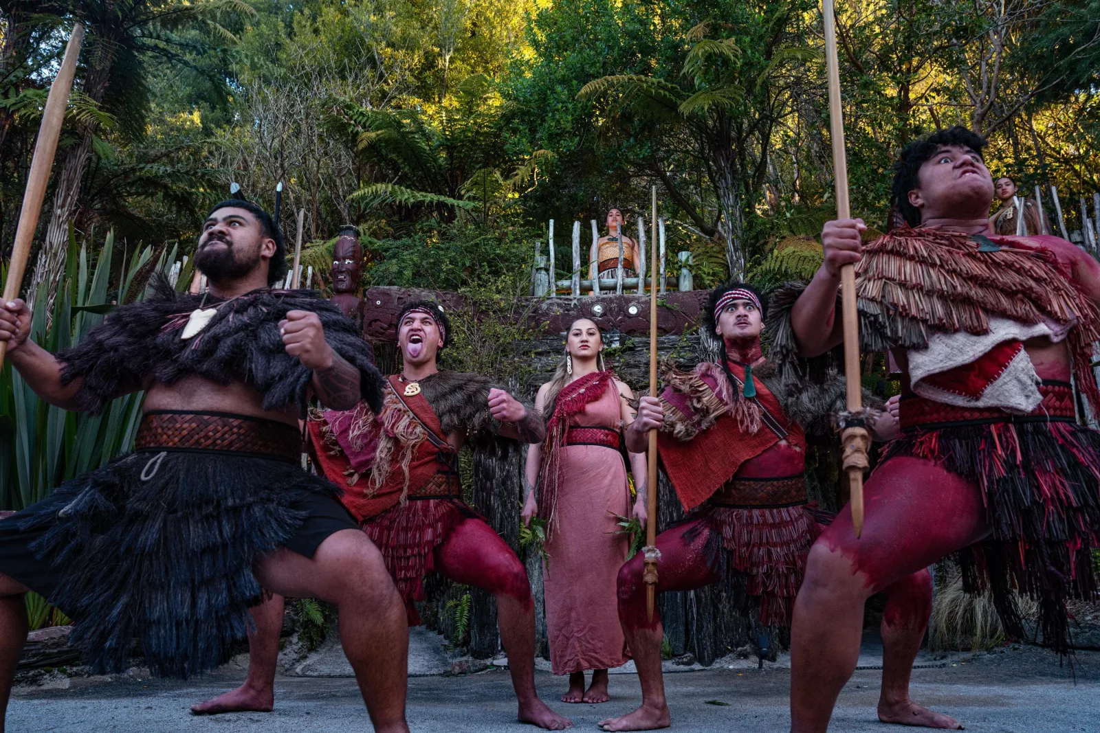 A line of Te Pā Tū performers with taiaha at dusk, a carved figure and palisade behind them