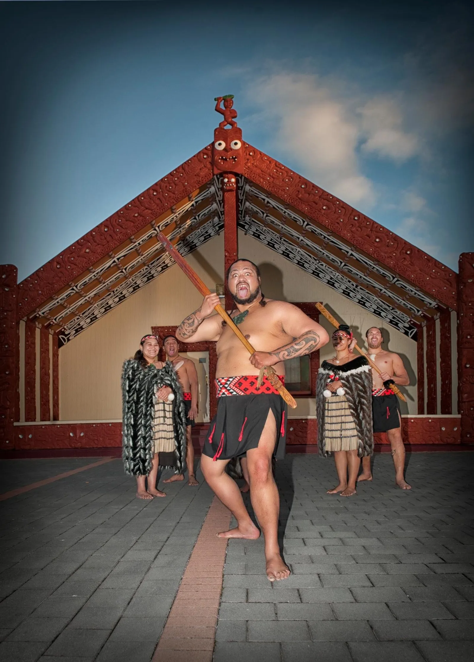 A Whakarewarewa performer with a taiaha in front of the carved meeting house, the rest of the group behind