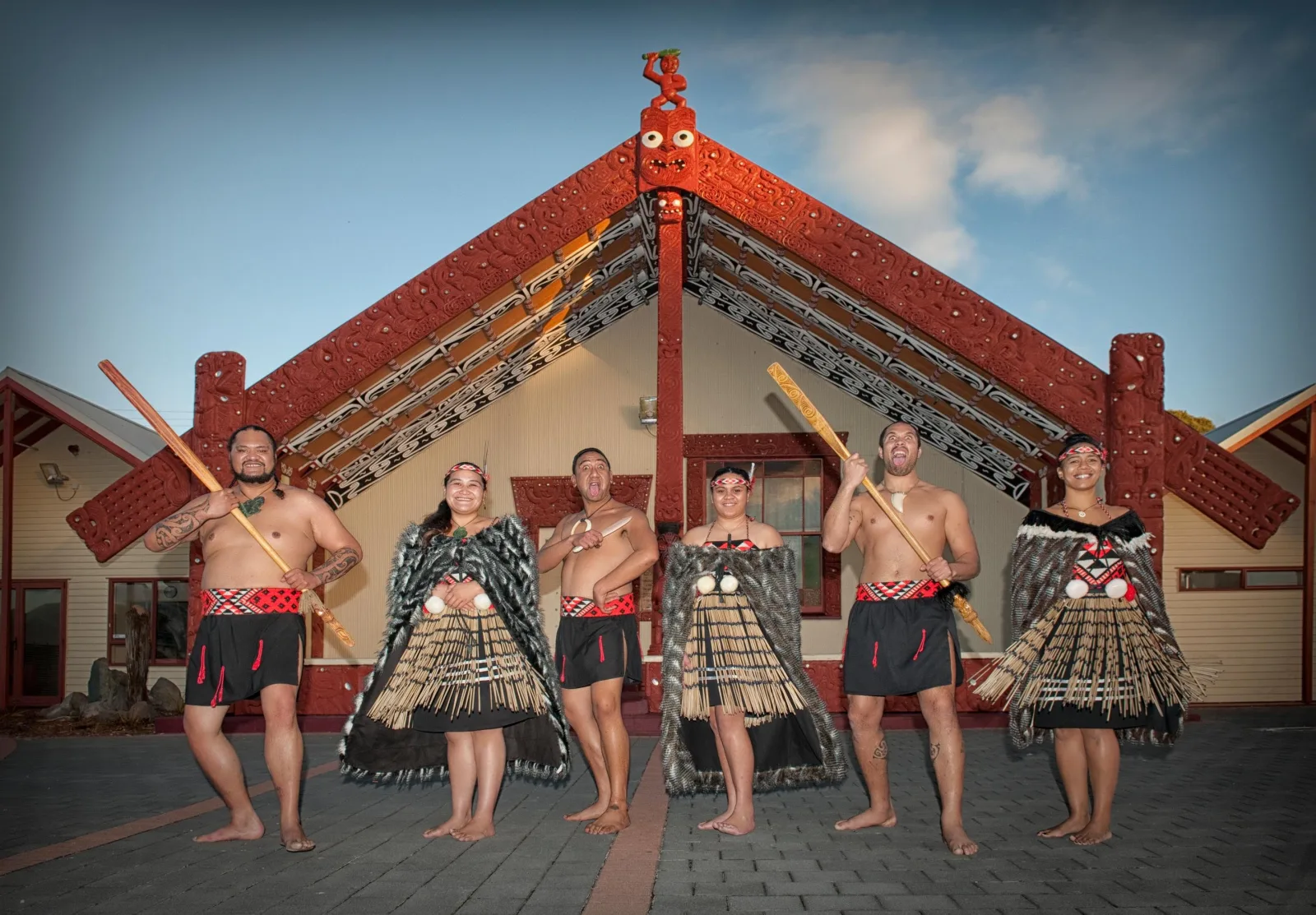 Six Whakarewarewa performers with hoe paddles in front of the carved meeting house at dusk