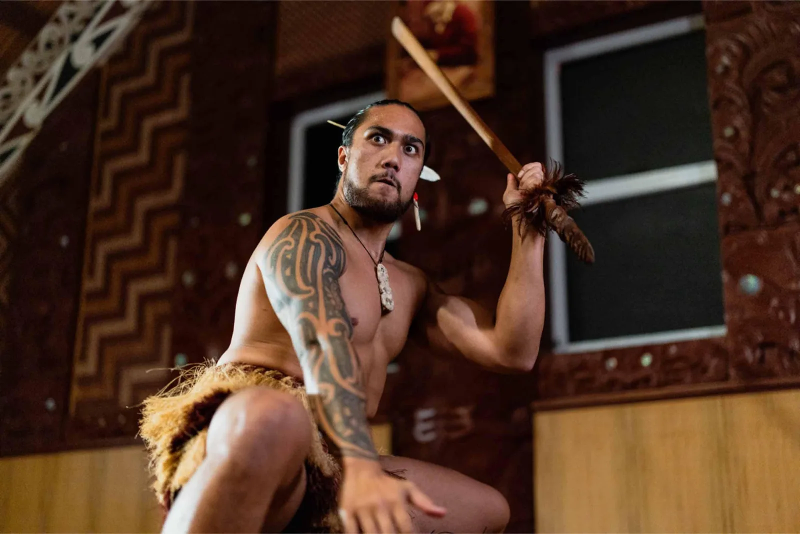 A Te Puia performer with a taiaha inside the carved meeting house, tukutuku panelling behind him