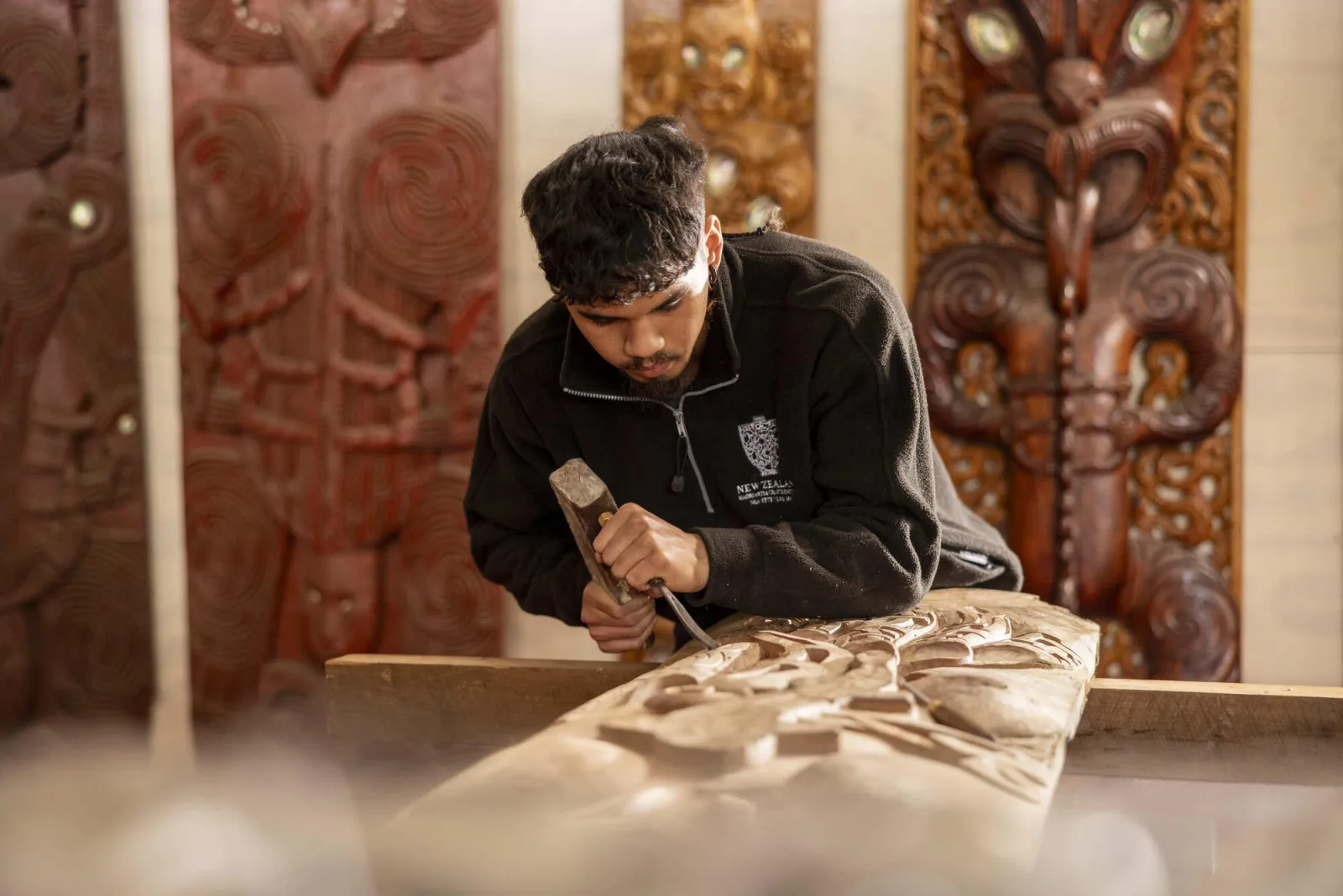 A carving student at the New Zealand Māori Arts and Crafts Institute working a panel with mallet and chisel