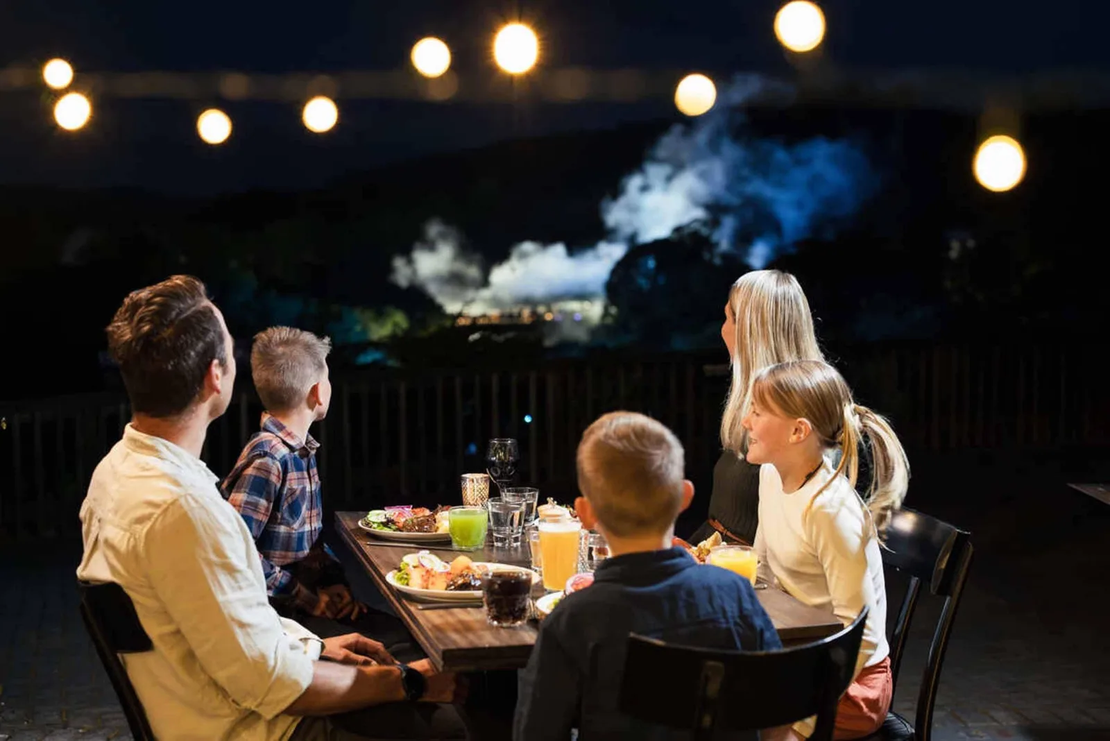 A family at dinner under festoon lights at Te Puia at night, a steam plume rising in the dark behind